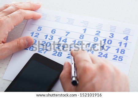Close-up Of Hand Holding Pen Over Calendar With Mobile Phone