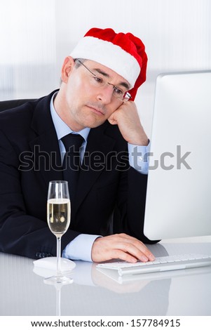 Sad Businessman Wearing Santa Hat Sitting In Front Of  Computer With Champagne Glass