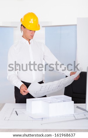 Male Architect Analyzing Blueprint With Architectural Model On Desk