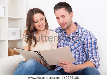 Young Couple Sitting Side By Side On Couch Looking At Photo Album