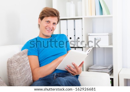 Young Man At Home Sitting On Couch Reading Book