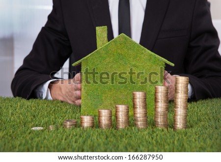 Businessman Holding Eco Friendly House In Front Of Stack Of Coins Over Grass