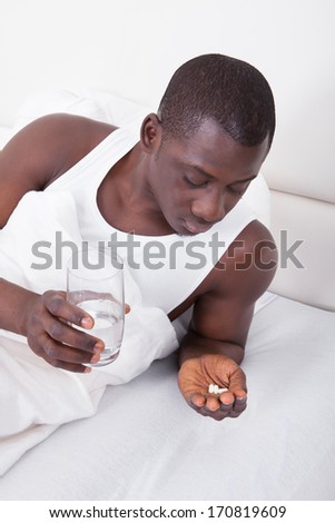 Young African Man Lying On Bed Holding Glass Of Water And Medicine