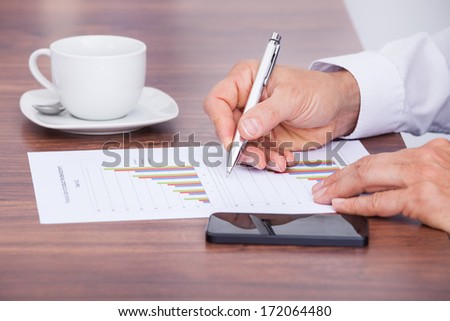 Businessman Writing On Document With Cup And Cellphone On Desk