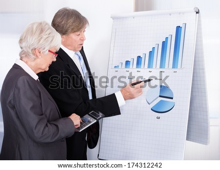 Mature Businessman And Businesswoman Calculating With Calculator In Front Of Chart On Flipchart