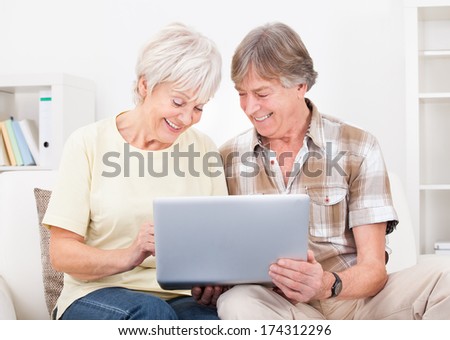Portrait Of Happy Senior Couple Sitting On Couch Using Laptop