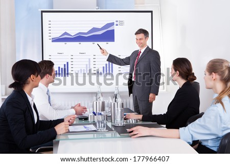 Mature Businessman Explaining Graph To His Colleagues Sitting In Office