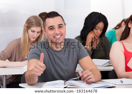 Portrait of confident male college student sitting at desk with classmates in background
