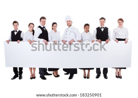 Large group of catering staff holding a blank banner with copyspace for your text including a chef waiters waitresses hostess and wine steward isolated on white