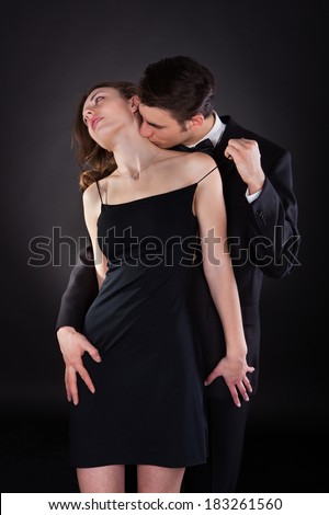 Young man in suit kissing woman on neck while removing dress strap from her shoulder isolated over black background