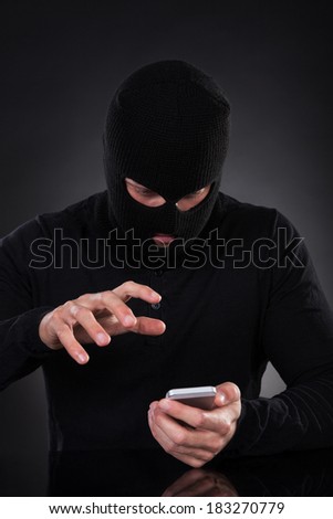 Thief in a balaclava and black outfit standing in the darkness trying to access a stolen mobile phone or a terrorist activating a bomb remotely