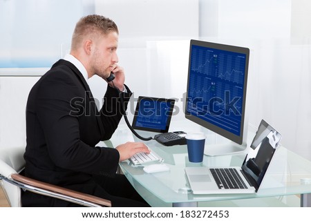 Hardworking businessman at his desk working on the computer screens simultaneously as he takes a call on his mobile phone
