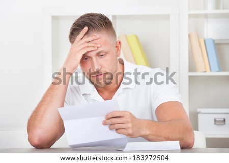 Businessman sitting in an office reacting in shock to the contents of a letter that he is reading raising his hand to his mouth