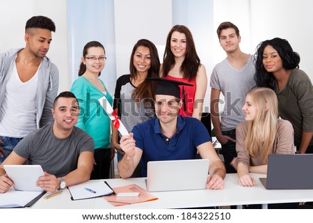 Portrait of college student holding degree with classmates looking at him in classroom