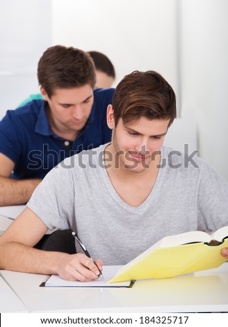 Young male university student writing while looking in book at desk in classroom