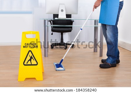 Yellow warning notice to caution people to a slippery wet surface as a janitor mops the floor in an office building Yellow warning notice to caution people to a slippery wet surface as a janitor mops the floor in an office building