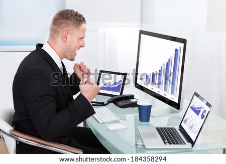 Businessman celebrating a performance graph sitting at his desk with statistical reports spread over three monitors cheering and clasping his fists
