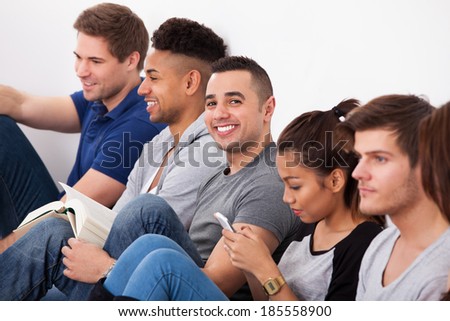 Portrait of happy male college student sitting with classmates against wall in classroom