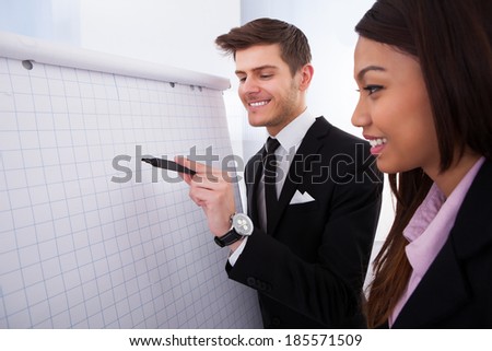 Businessman writing on flipchart with female colleague standing by him in office