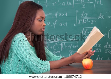 Side view of African American teacher reading book at classroom desk