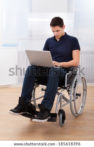 Full length of young handicapped man using laptop while sitting on wheelchair at home