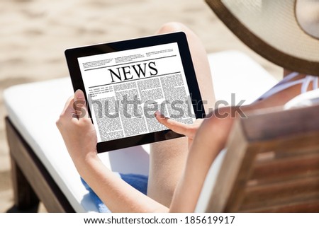 Woman reading newspaper on digital tablet while relaxing on deck chair at beach