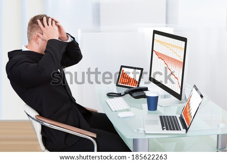 Despairing businessman faced with financial losses sitting at his desk consulting three graphs on different monitors all dropping into the red