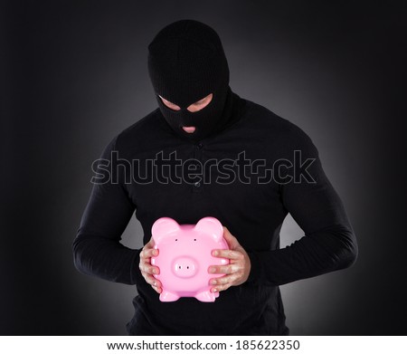 Furtive thief or burglar wearing a black outfit and balaclava making his escape through darkness stealing a pink piggy bank conceptual of theft of savings and investments