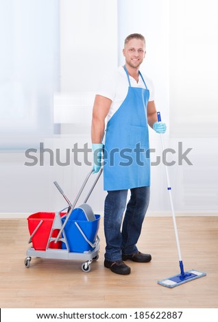 Janitor cleaning wooden floors with a mop and a cart with two buckets for the disinfectant and water pausing to smile at the camera as he goes about his work in an office building