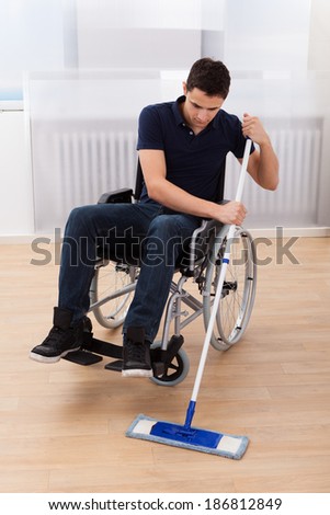 Full length of young handicapped man mopping hardwood floor while sitting on wheelchair in house