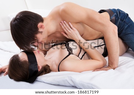 Side view of young man kissing sexy blindfolded woman in bed at home
