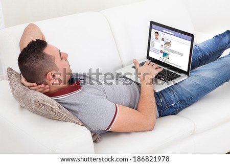 Young man chatting on laptop while lying on sofa at home