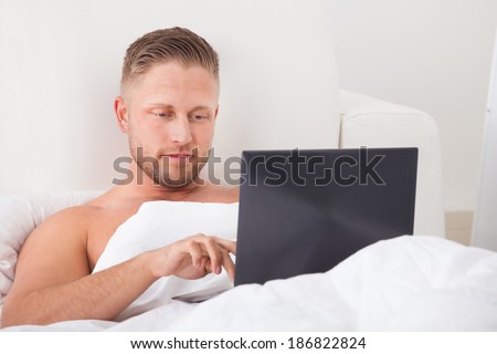 Man sitting up in bed against the pillows working on a laptop computer smiling as he reads the screen