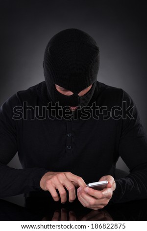 Thief in a balaclava and black outfit standing in the darkness trying to access a stolen mobile phone or a terrorist activating a bomb remotely