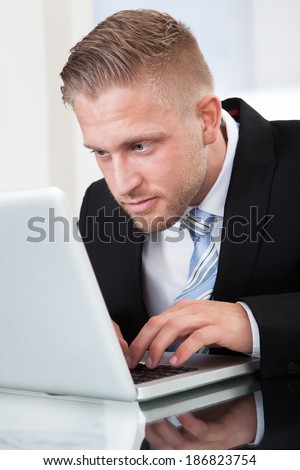 Businessman leaning forward staring intently at his laptop screen as he types information on the keyboard while sitting at his desk  close up portrait