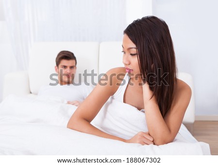 Depressed young woman sitting on mattress with angry man in background