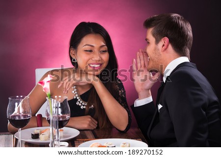 Happy young man whispering in woman\'s ear at restaurant table