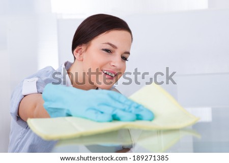Smiling young maid cleaning glass table at home