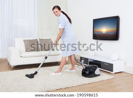 Full length portrait of young maid cleaning carpet with vacuum cleaner at home