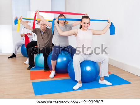 Portrait of trainer and senior customers with resistance bands sitting on fitness balls at gym