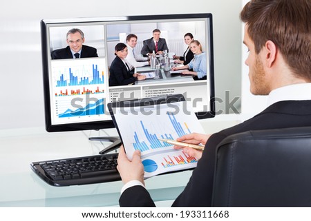 Young businessman analyzing graphs while video conferencing with colleagues on computer at office desk