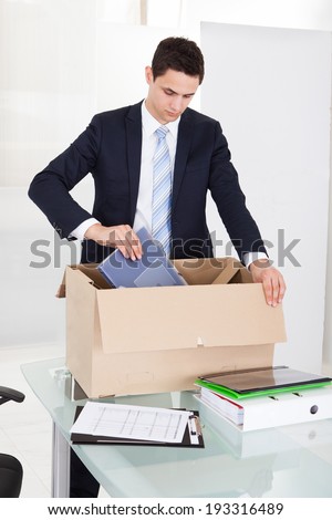 Sad young businessman packing files in cardboard box at desk in office