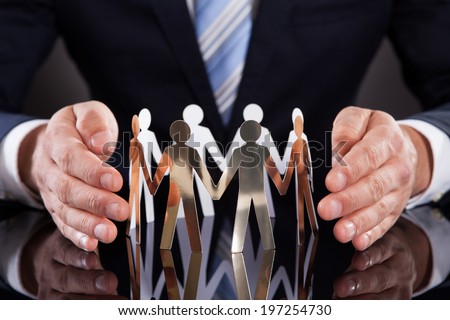 Midsection of businessman\'s hands protecting team of paper people on desk against black background