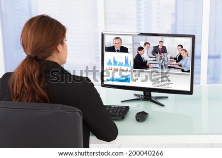 Rear view of businesswoman video conferencing with team on computer at desk in office