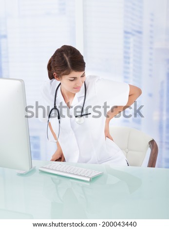 Sad female doctor with backache at computer desk in clinic