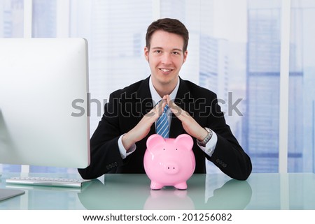 Midsection of young businessman protecting piggy bank at office desk