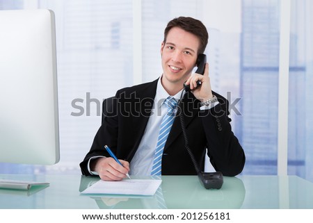 Portrait of young businessman using telephone while writing on document at office desk
