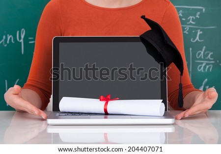 Midsection of young college student showing laptop with mortarboard and degree in classroom