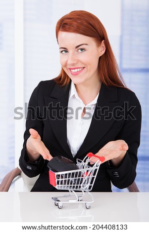 Portrait of confident businesswoman showing shopping cart model with computer mouse on desk in office