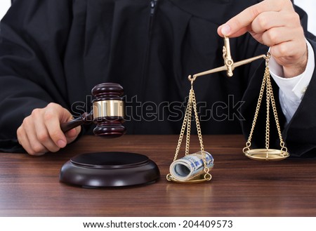 Midsection of male judge striking gavel while holding scale with money in courtroom